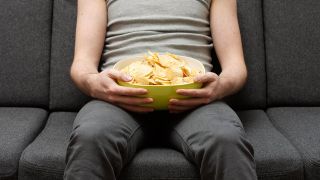 Ein Mann sitzt auf dem Sofa und hÃ¤lt eine SchÃ¼ssel mit Chips auf dem SchoÃ. 