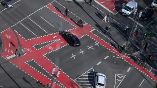 Radfahrer, FuÃgÃ¤nger und Autos sind an einer Kreuzung, die mit roten Fahrradspuren markiert ist, unterwegs. 