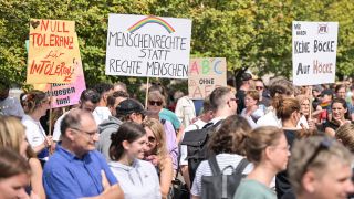 Demonstranten halten mehrere Schilder hoch, unter anderem mit der Aufschrift 