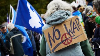 Zur Friedensdemonstration versammeln sich einige Menschen. Eine Frau trägt ein Schild mit der Aufschrift "No War".