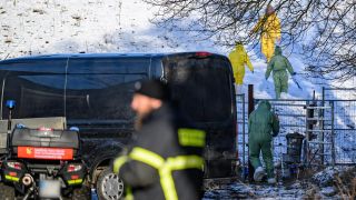 Personen in Schutzkleidung gehen über eine Weide, während Feuerwehr und Polizei das Gelände weiträumig abgesperrt haben.