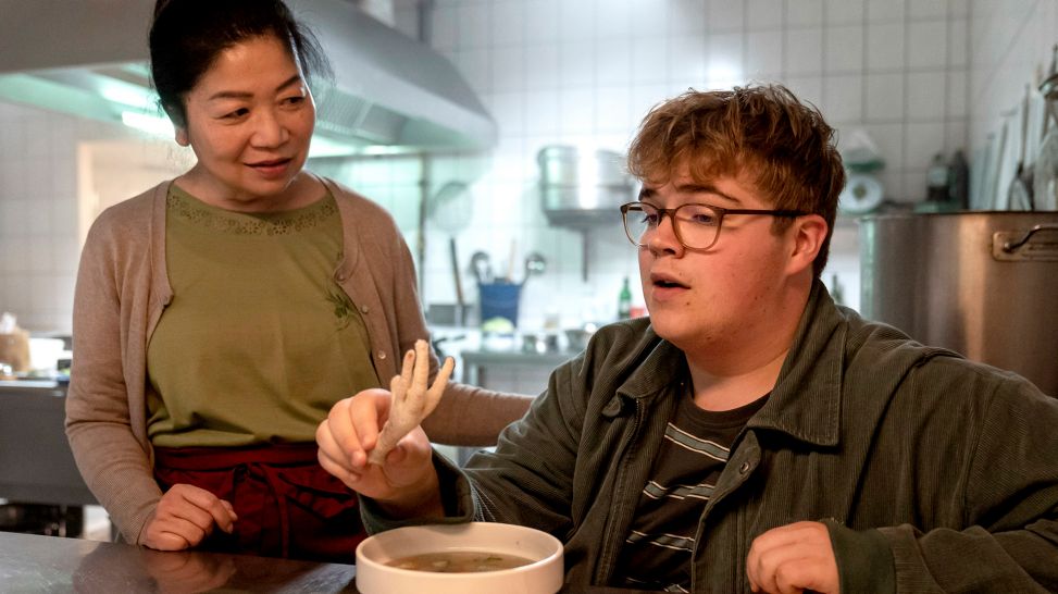 Jannik (Lorenzo Germeno) beeindruckt Tais Großmutter Linh (Mai Phuong Kollath), weil er auch vor Hühnerfüßen nicht zurückschreckt; Szene aus der Serie "Nackt über Berlin"; Quelle: SWR/Studio.tv.film/Oliver Feist