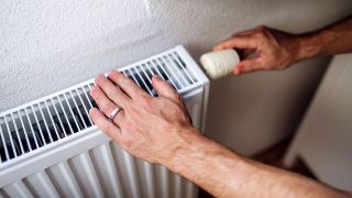 Ein Mann dreht das Thermostat an einem Heizkörper und fasst ihn dabei mit der anderen Hand an (Quelle: IMAGO / Westend61)