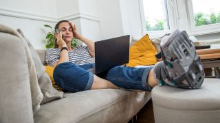 Eine Frau sitzt mit gebrochenem Fuß und Laptop auf dem Schoß auf einem Sofa und telefoniert (Quelle: picture alliance / dpa-tmn | Christin Klose)